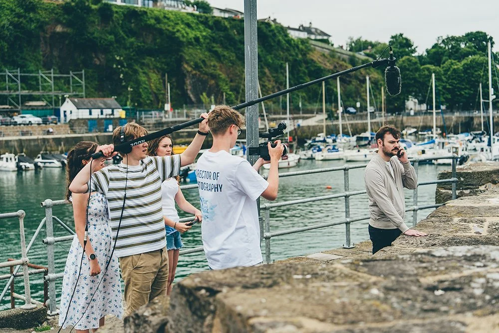 Film crew filming a man by the harbor, with boats and hills in the background.