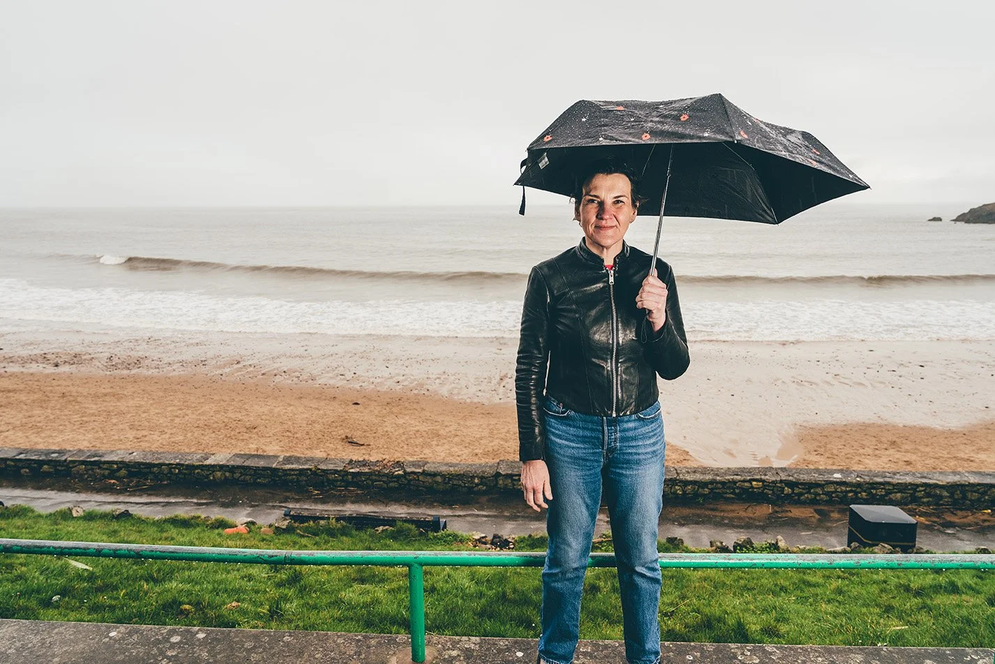 A woman standing outdoors on a beachside promenade holding a black umbrella, wearing a black leather jacket and blue jeans, with an overcast sky and ocean in the background.