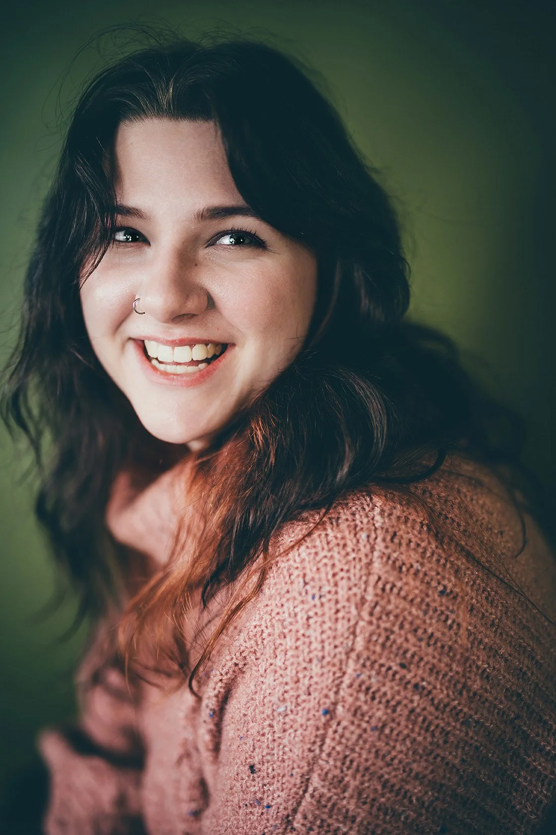 A woman with dark wavy hair and a nose ring smiling in front of a green background.