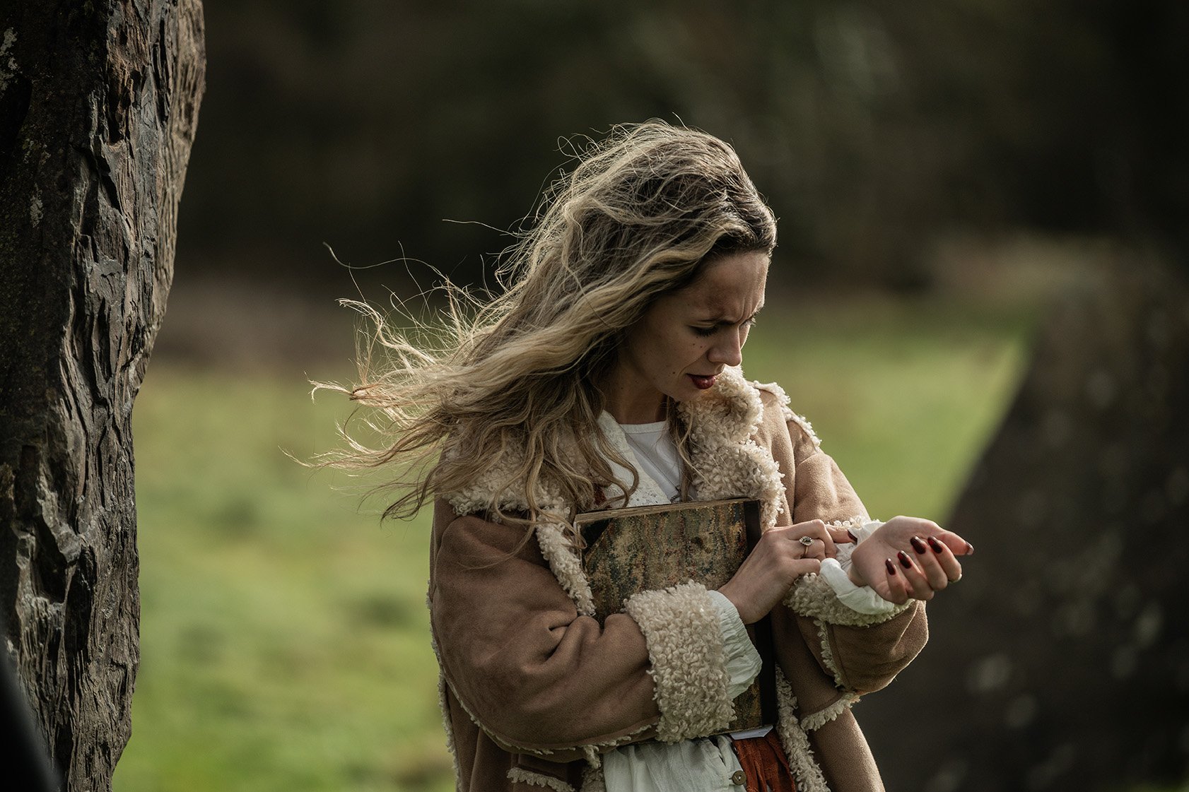 A woman with long, wavy blonde hair standing outdoors during daytime, holding a book and looking at her wrist with a concerned or curious expression.