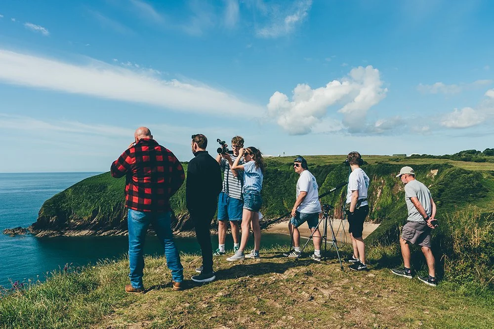 Group of people outdoors on a grassy cliff overlooking the ocean, some taking pictures and observing the view on a partly cloudy day.