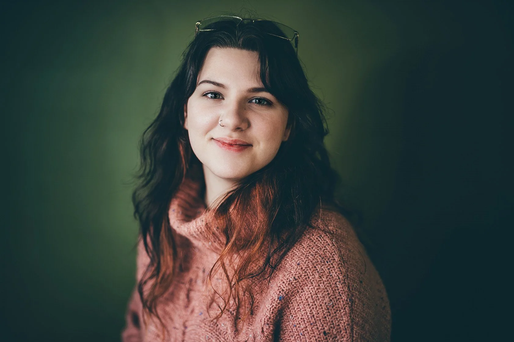 A young woman with dark wavy hair, wearing glasses on her head, a nose ring, and a pink cozy sweater, smiling at the camera against a dark green background.