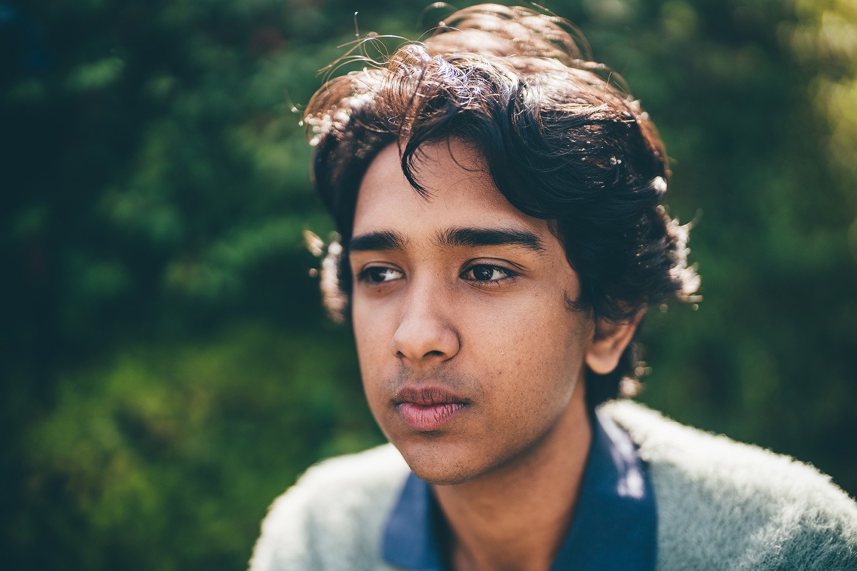 Close-up of a young man with wavy dark hair and tan skin, looking contemplative outdoors with a blurred green background.