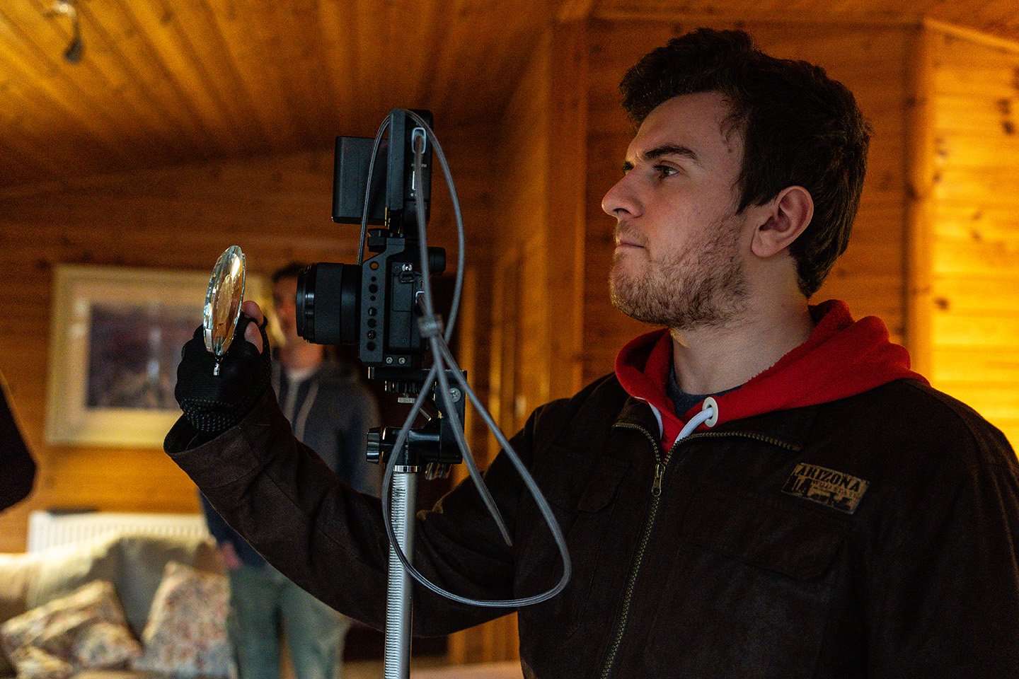 A young man with dark hair, beard, and wearing a black jacket with a patch, is holding a camera with a large lens, focusing intently on a shot inside a wooden cabin.