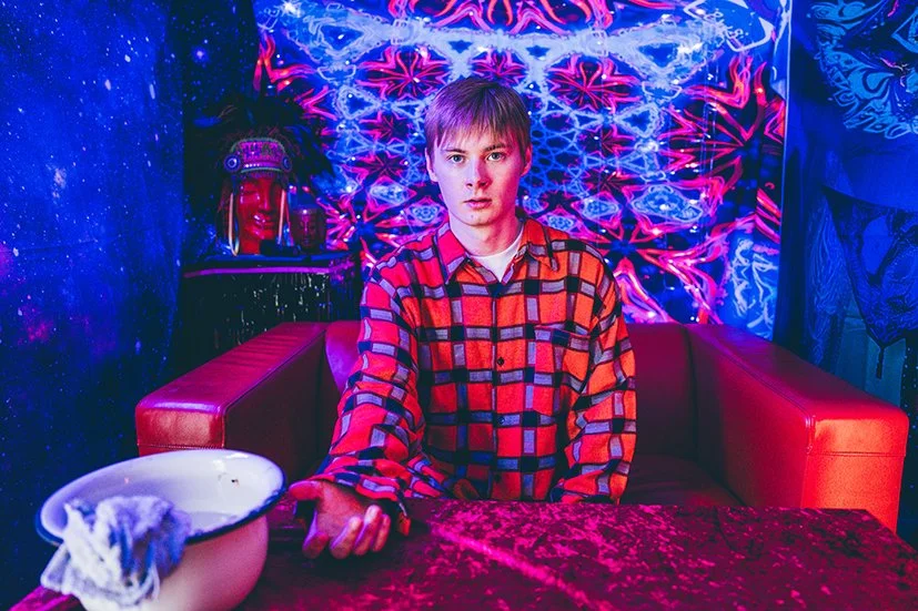 Young man in red and black plaid shirt sitting at a table with a white bowl and cloth, in a room with vibrant, psychedelic wall art and glowing decorations.