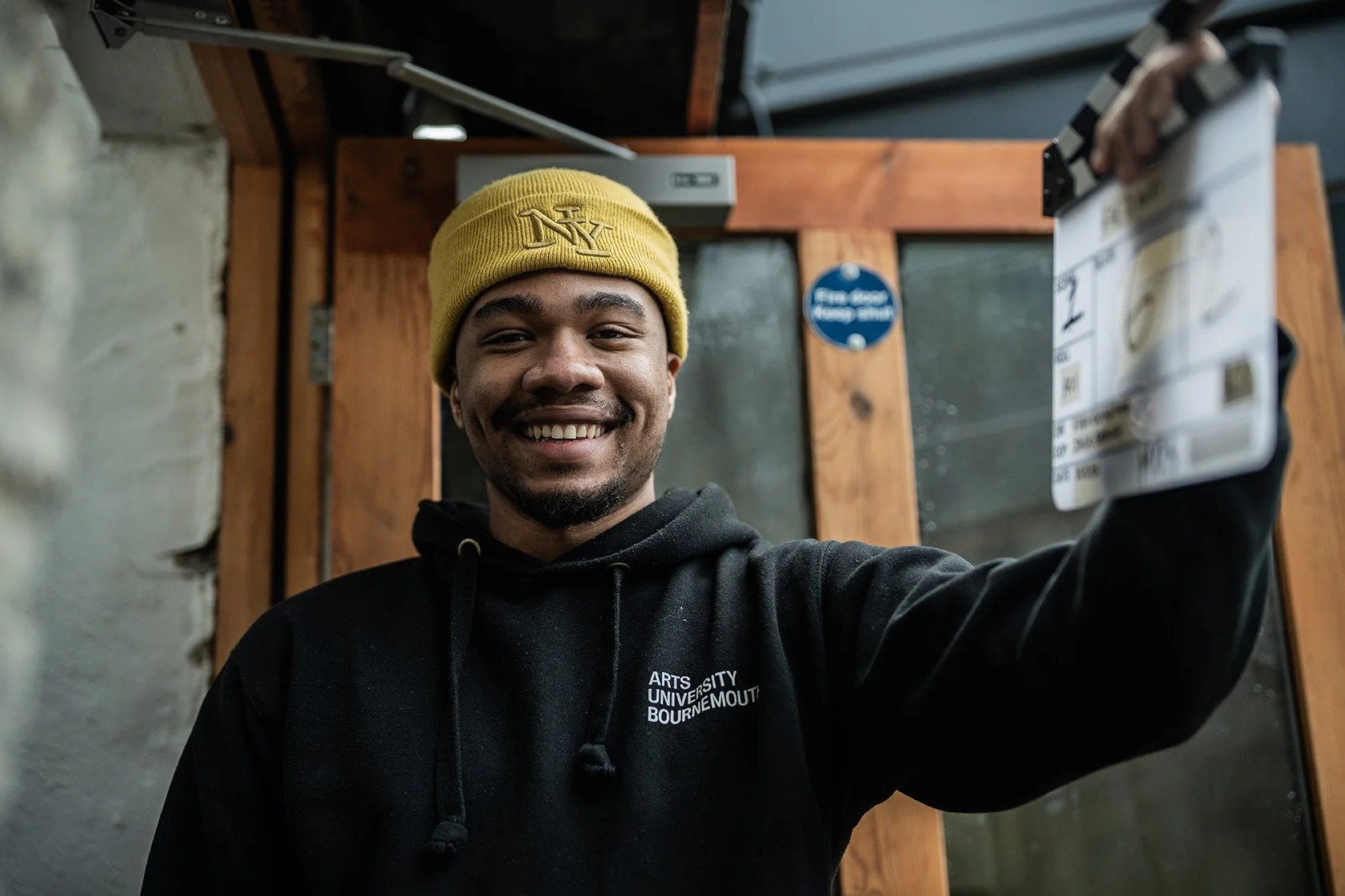 A smiling young man wearing a yellow beanie and a black hoodie with white text, holding a bus or train ticket while standing indoors near a wooden door with a glass window.
