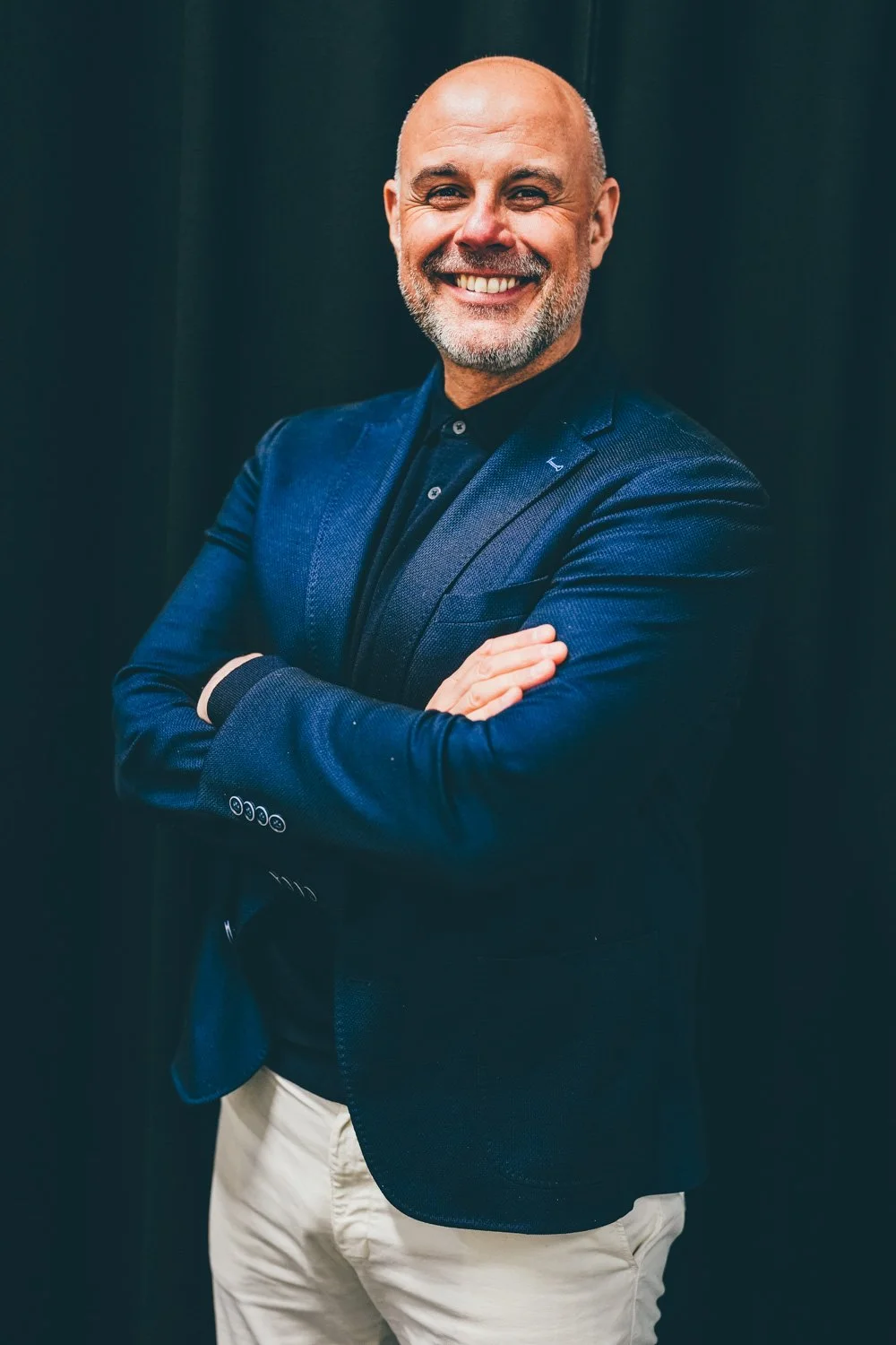 A smiling, bald man with a beard wearing a dark blue blazer and light-colored pants, standing with arms crossed against a dark background.