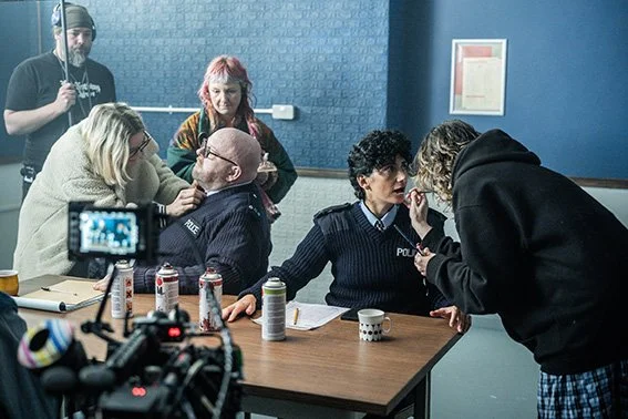 Filming scene with actors and crew; woman in black sweater talking to police officer, other cast members and crew in the background.