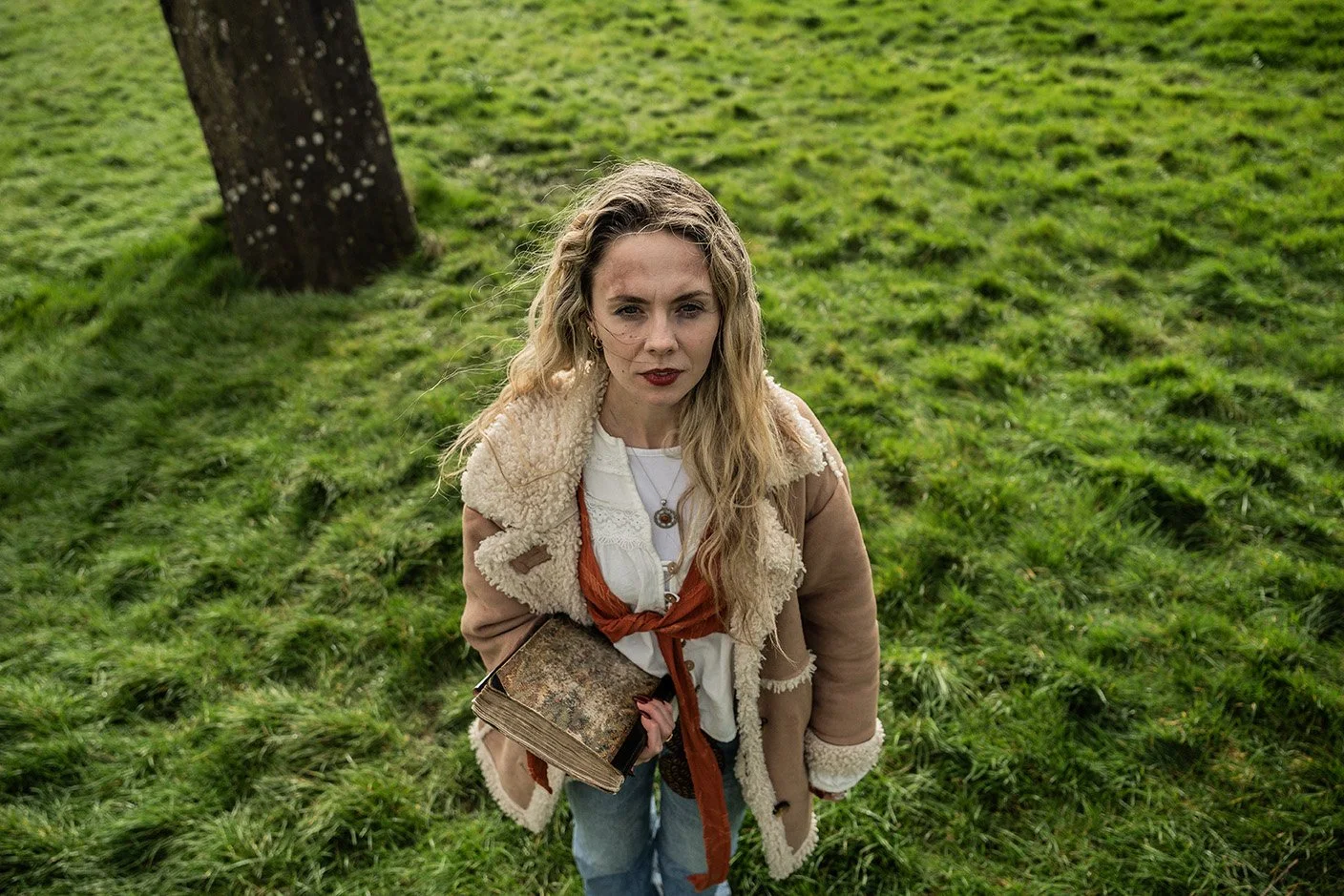 A woman with long wavy blonde hair and red lipstick looking up at the camera, standing outdoors on grassy ground near a tree, holding a large book, wearing a tan shearling coat over a white shirt