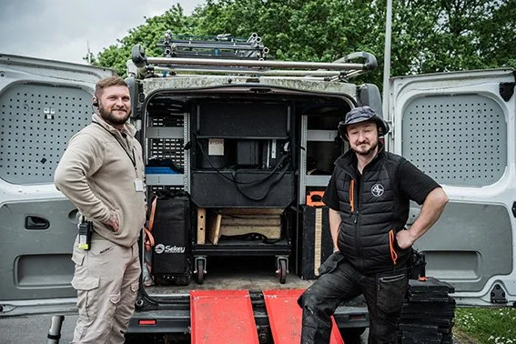 Two men standing in front of an open mobile equipment truck, with various gear inside, outdoors on a cloudy day.