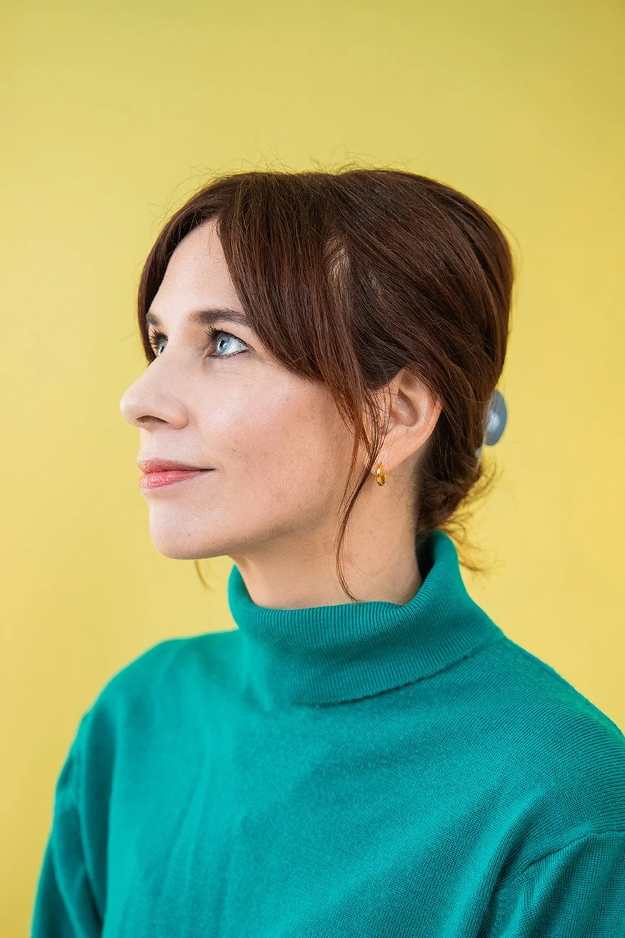 Side profile of a woman with auburn hair and blue eyes wearing a teal turtleneck sweater against a yellow background.