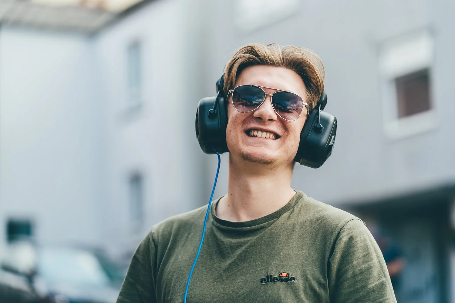 A young man with short, light brown hair wearing sunglasses and large black headphones, smiling outdoors in front of a light-colored building.