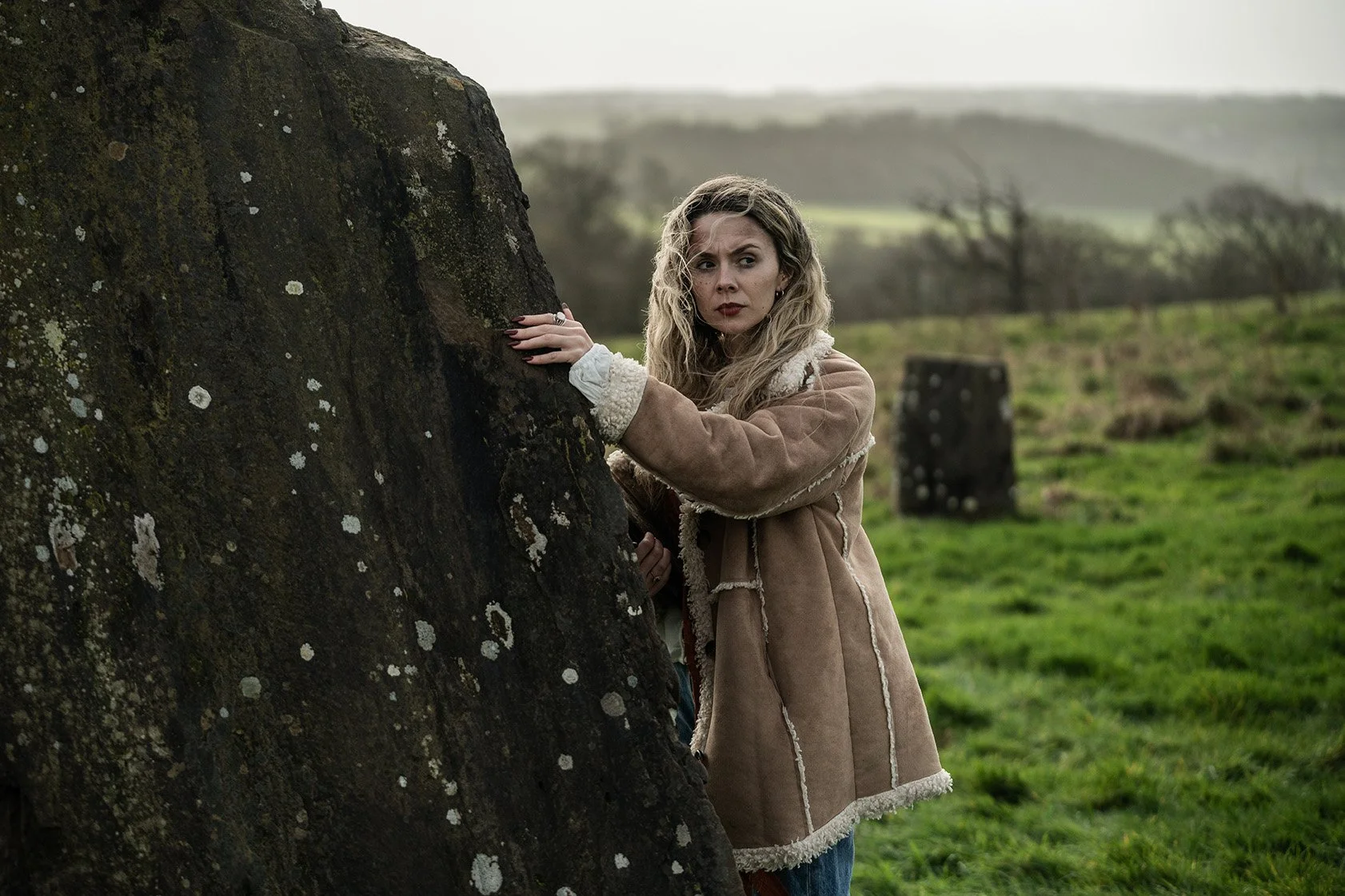A woman with blonde curly hair wearing a tan shearling coat is standing behind and holding onto a large tree trunk in a grassy field, looking sideways with a concerned or cautious expression, with rolling hills and trees in the background.