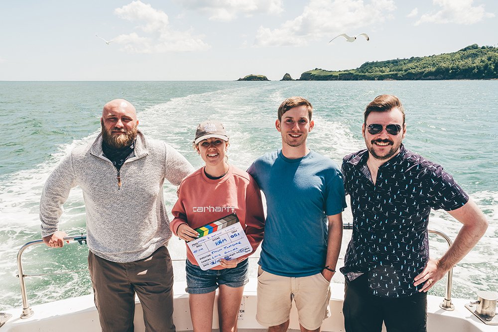 Group of four friends smiling on a boat, with water and a distant shoreline in the background, and a seagull flying overhead.