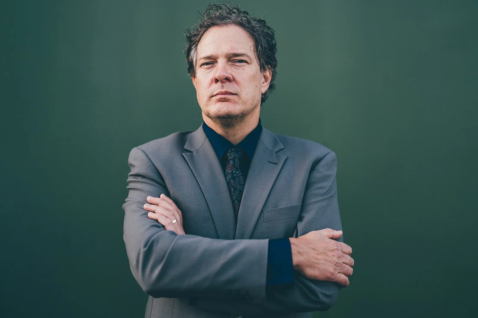 A man with curly dark hair, wearing a gray suit, dark shirt, and patterned tie, standing with crossed arms in front of a dark green background.