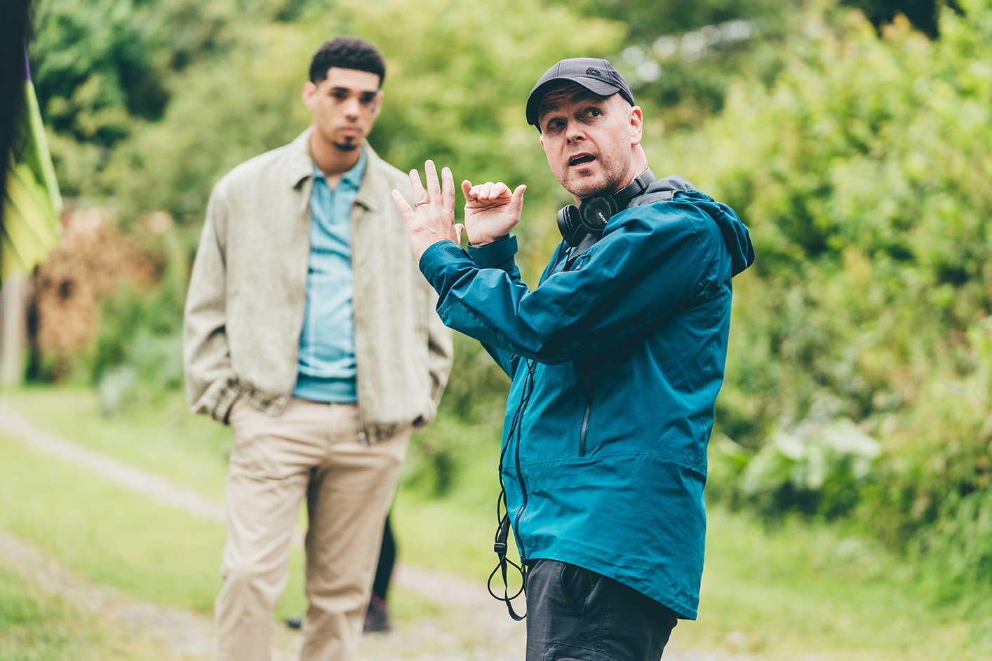 A man in a blue jacket and black cap gives direction or instructions while another man in a beige jacket and light-colored pants listens, with greenery in the background.
