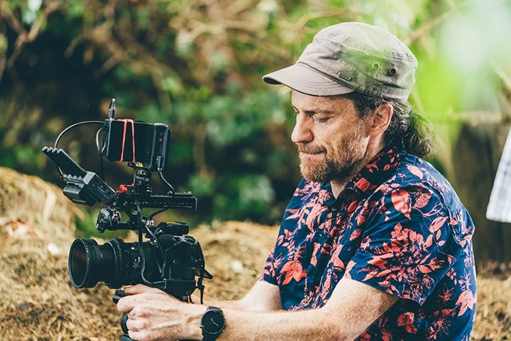 A man in a floral shirt and gray hat operating a professional video camera outdoors surrounded by greenery.