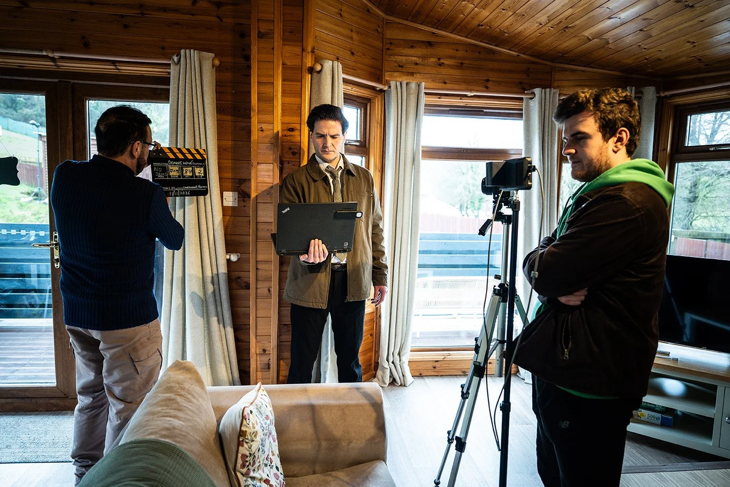 Three men working on a film or video shoot inside a wooden-paneled living room with large windows and curtains.