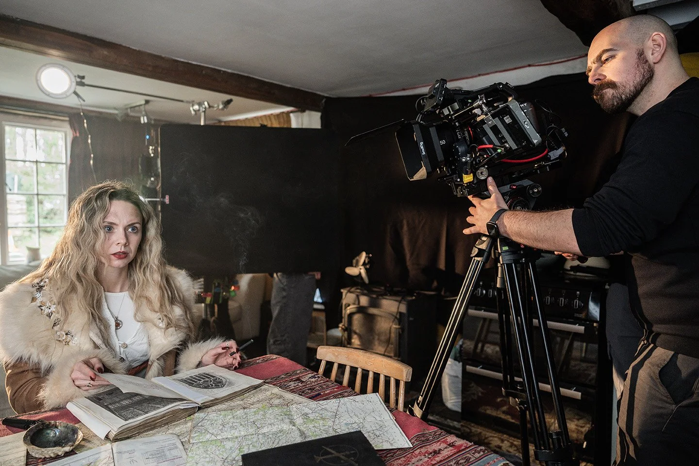A woman with long blonde, curly hair and a beige fur coat is sitting at a wooden table with newspapers, a map, and a bowl. A man with a shaved head and beard is operating a professional video camera on a tripod, filming her inside a room with a windo