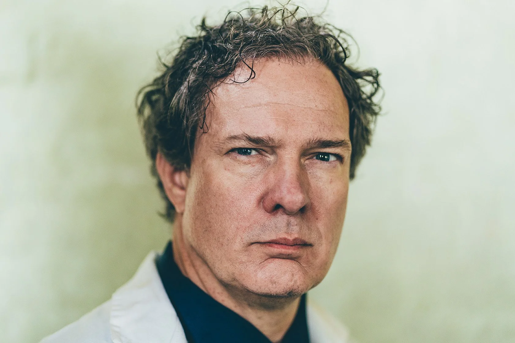 Close-up of a man's face with curly brown hair, blue eyes, and a serious expression, wearing a white coat and a dark shirt.