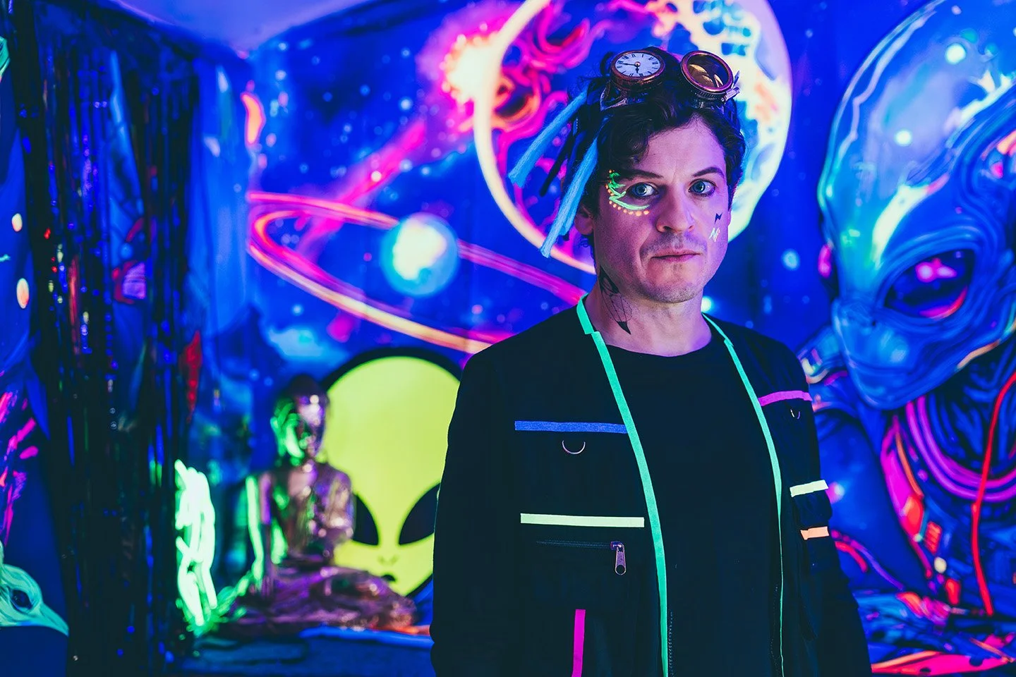 A person with tattoos and neon face paint, wearing goggles and a black jacket with colorful accents. They are in a vibrant, neon-lit art installation with psychedelic cosmic artwork, a radioactive symbol, and a figure sitting on the floor.