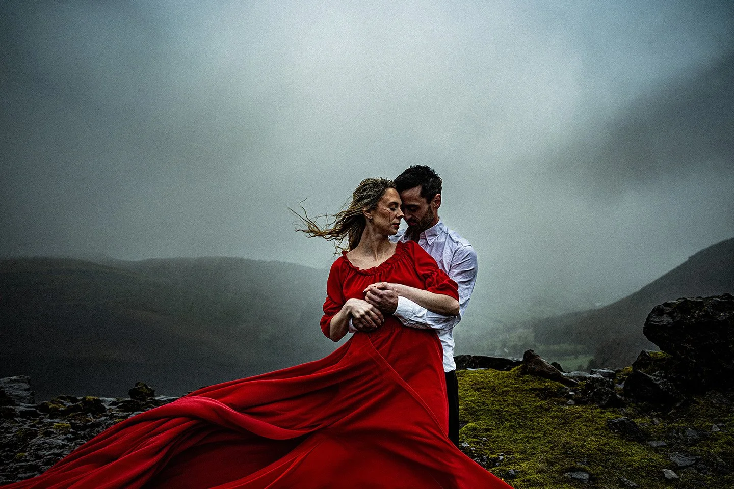 A couple standing outdoors on a rocky landscape under a cloudy sky, with the woman wearing a flowing red dress and the man in a white shirt, embracing and looking at each other.