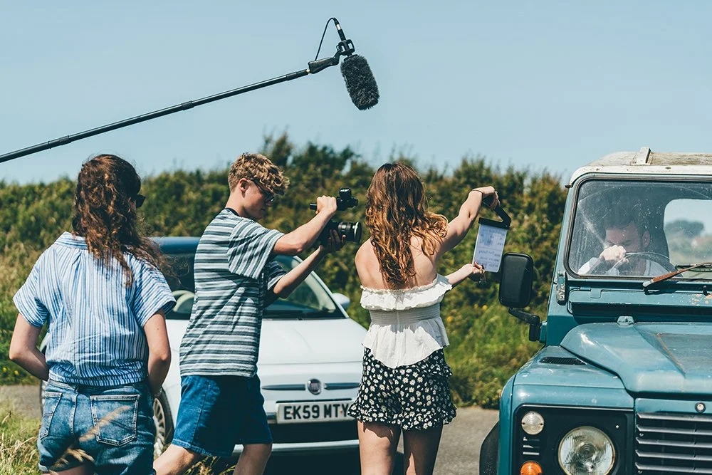 Filmmaking crew filming outdoors with a boom mic, camera, and a woman acting or speaking into a device next to a vintage jeep, with two other women observing.