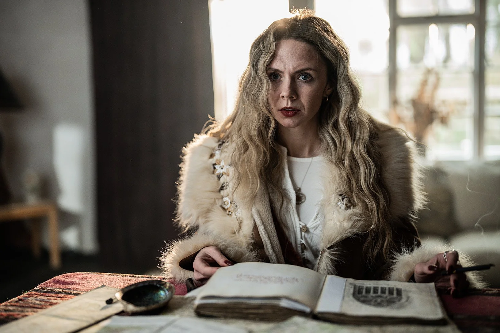 Woman with long wavy blonde hair, wearing a fur-lined jacket, sitting at a table with an open notebook, looking intently at something outside the frame.