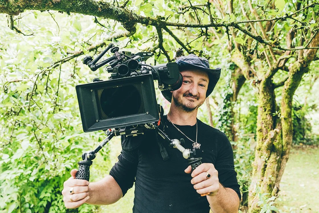 A man holding a professional video camera outdoors in a lush green, wooded area.