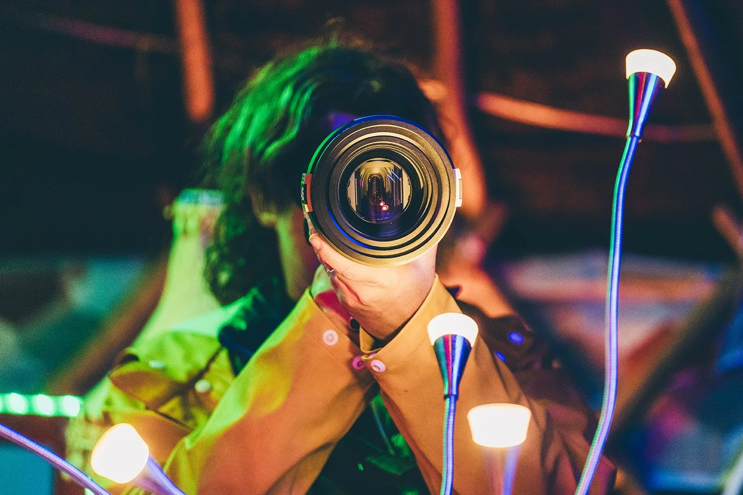Person holding a camera lens up to their face, with colorful lighting and decorative lights surrounding them in a dark setting.