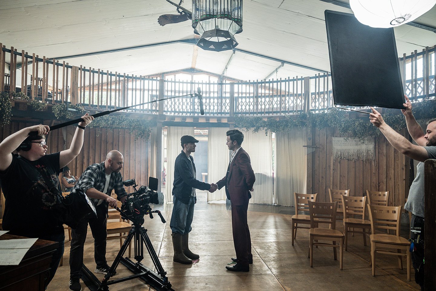 Two men in suits shake hands in the center of a wooden room while a film crew films the scene. Crew members hold equipment including a boom microphone and reflectors.