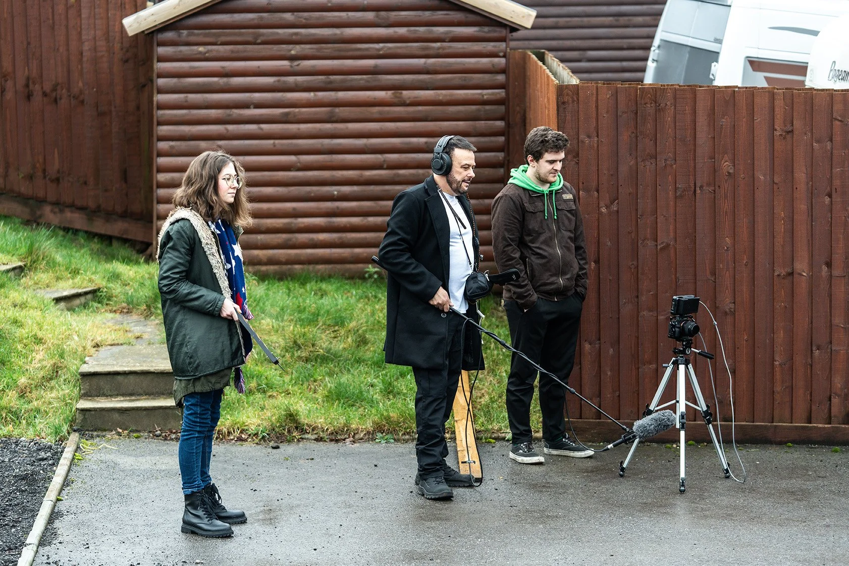 A group of three people filming outdoors with a camera on a tripod and a boom microphone, standing near a wooden fence and a small wooden shed.