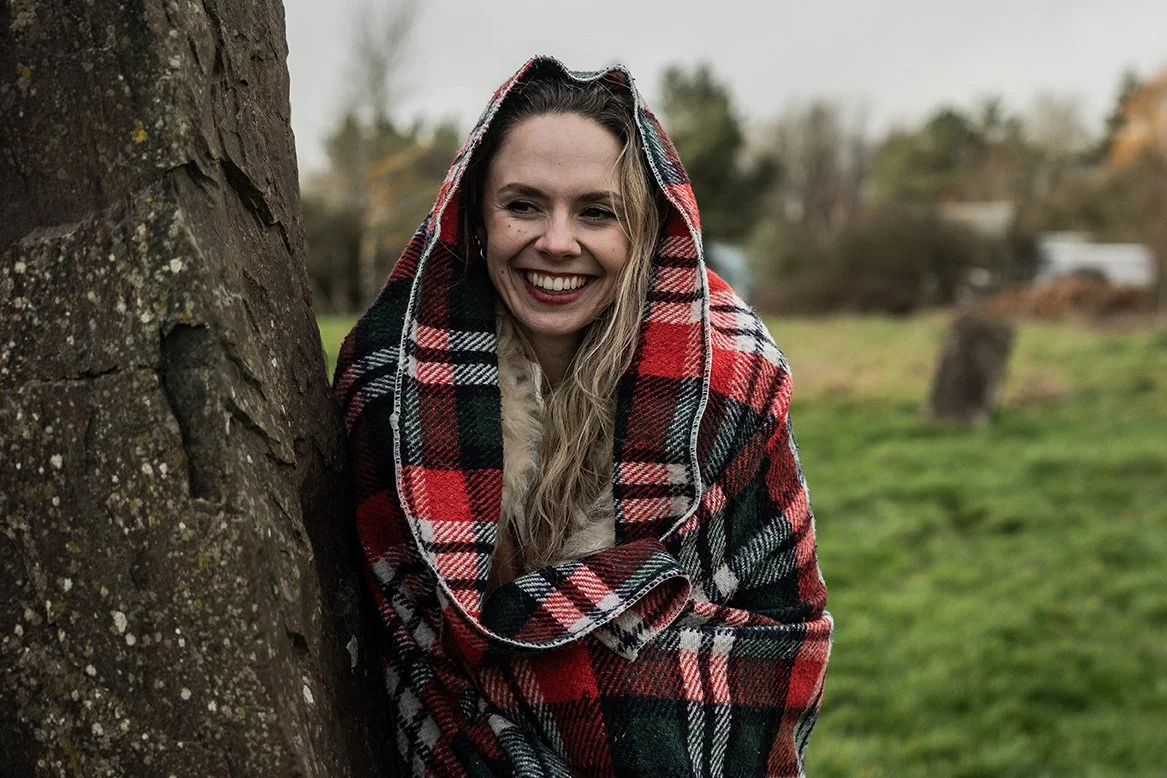 A smiling woman with long blonde hair wrapped in a red, black, and white plaid blanket outdoors near a tree, with a grassy field and trees in the background.