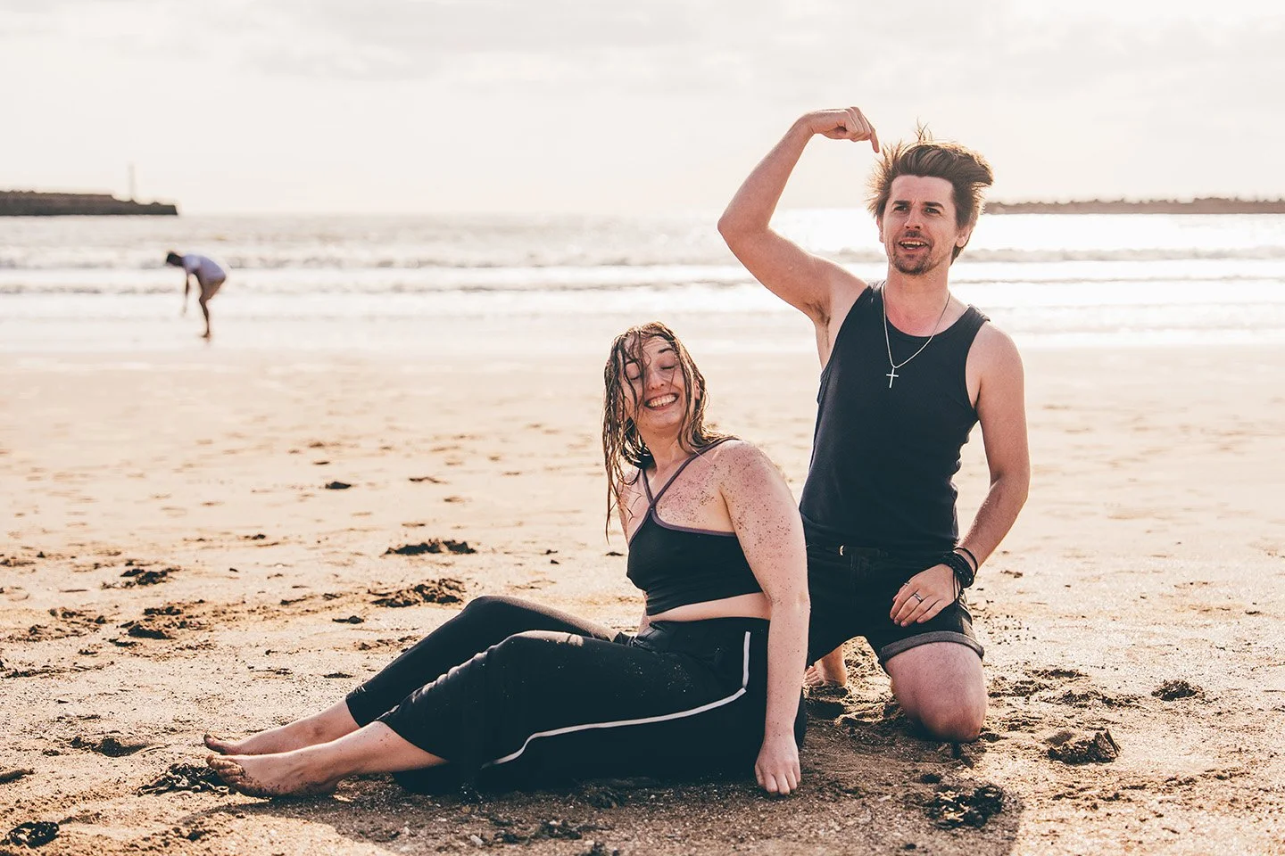A young woman and man sitting on the beach, smiling and enjoying the sunny day, with the ocean and a person wading in the background.