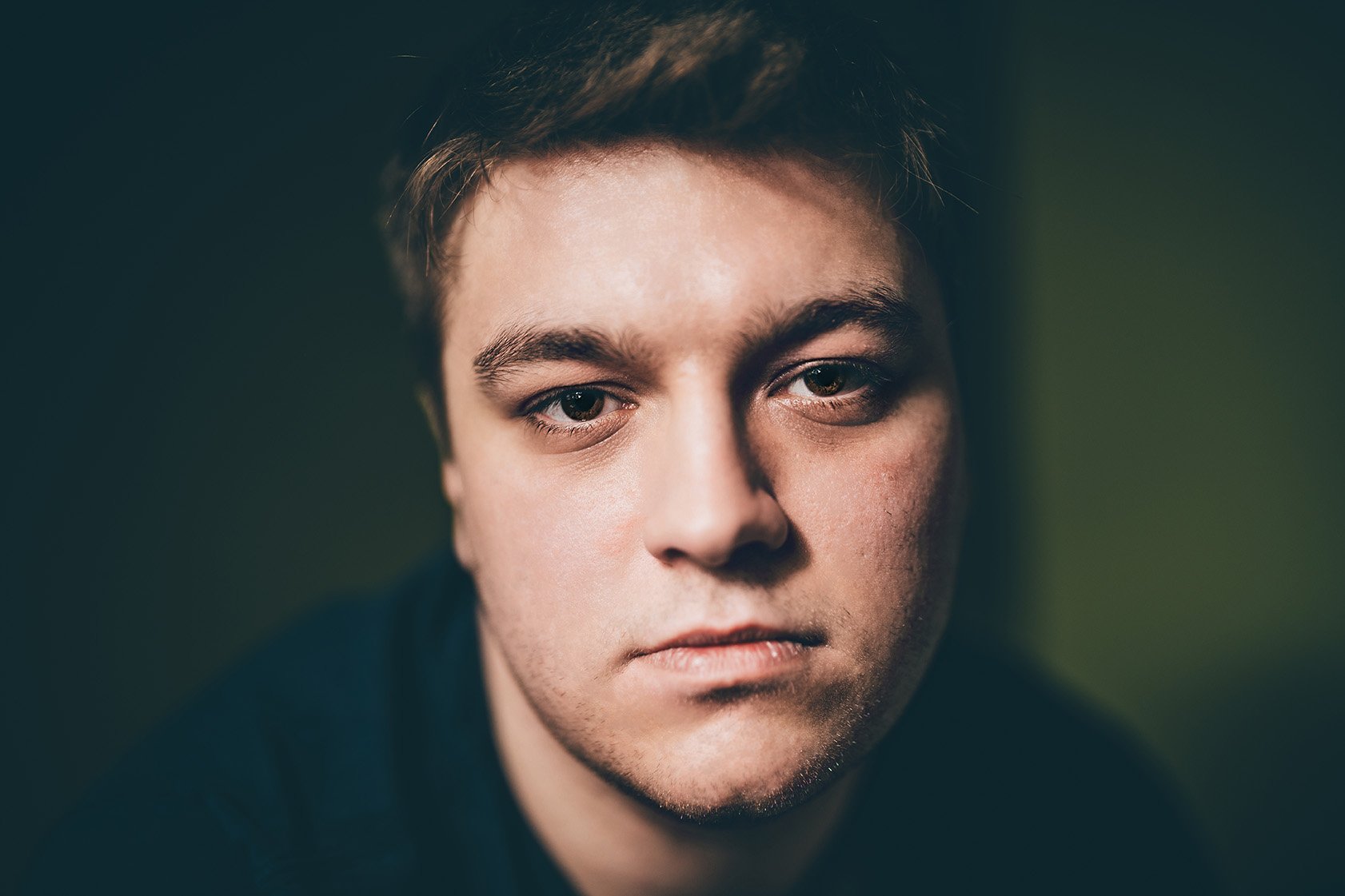 Close-up portrait of a young man with light skin, short brown hair, and brown eyes, looking directly at the camera with a serious expression.