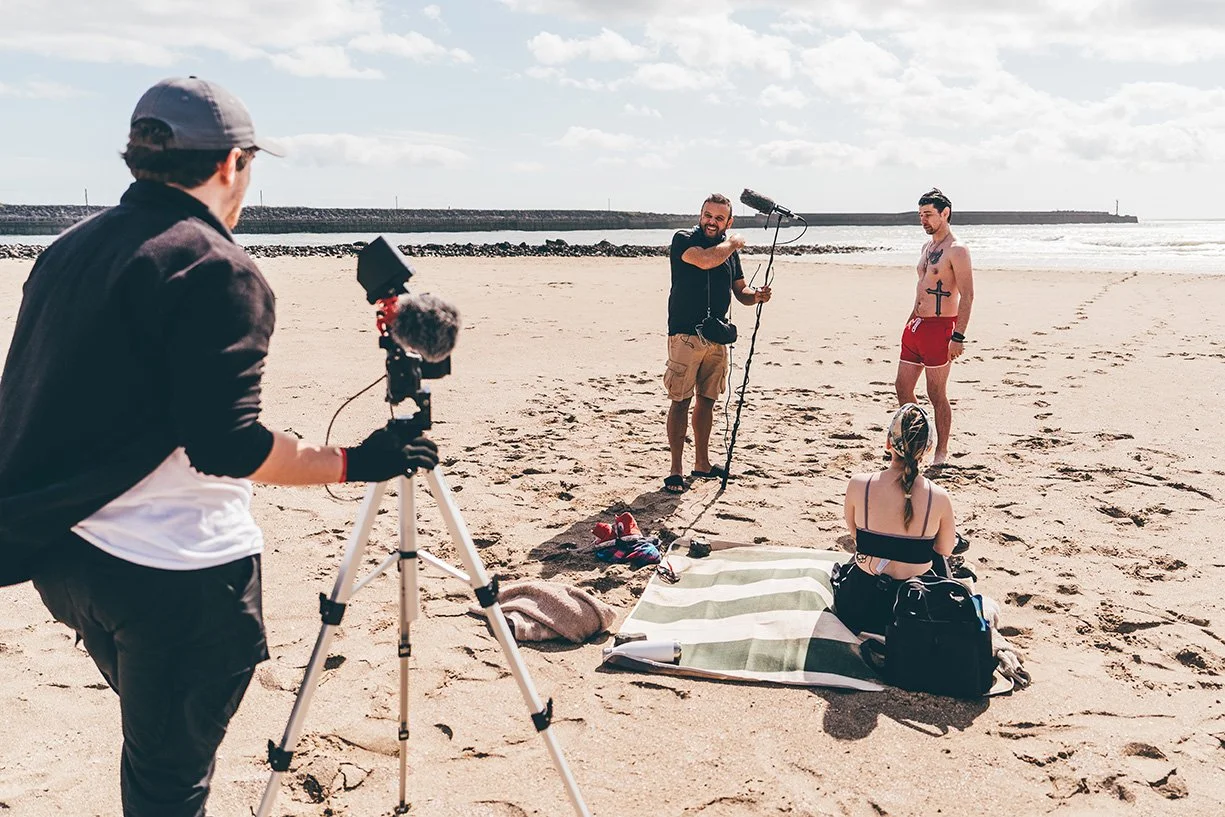 A beach scene with a video crew filming a man and a woman. The man with tattoos wears red shorts, and the woman sits on a striped towel. The crew includes a person operating a camera and another person holding a boom microphone. The background featur