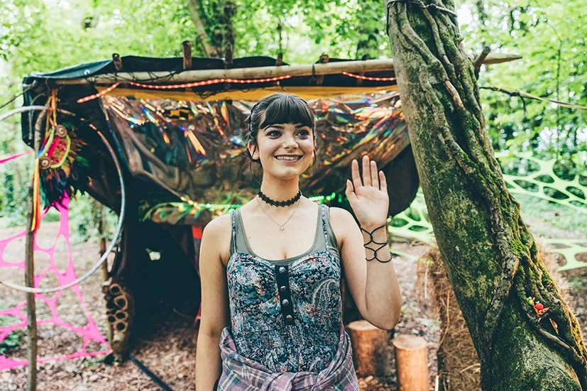 A young woman with dark hair and a choker necklace smiling and waving in a forest with a decorated, makeshift shelter in the background.