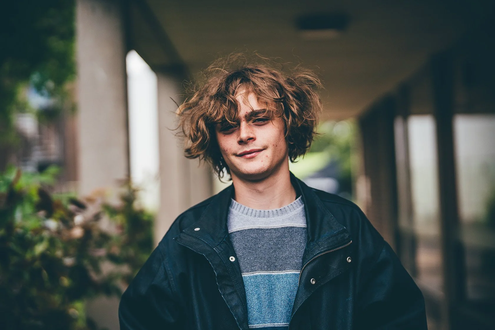 A young man with curly messy hair, wearing a striped sweater and black jacket, standing outdoors under an overhang with greenery in the background.