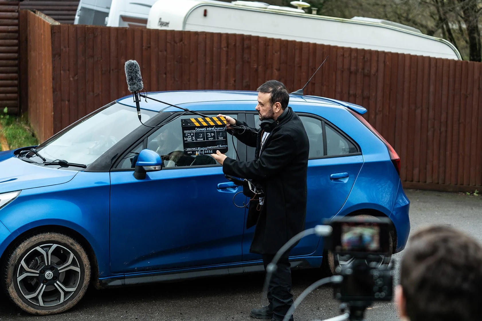 A man filming a scene in a blue car with a clapperboard on the window, during a film shoot, outdoors with a wooden fence and camper in the background.