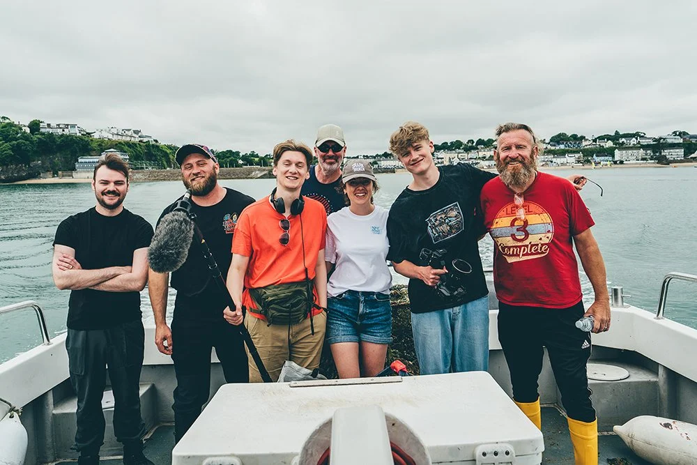 Group of seven people standing on a boat with a river and shoreline in the background, smiling at the camera.