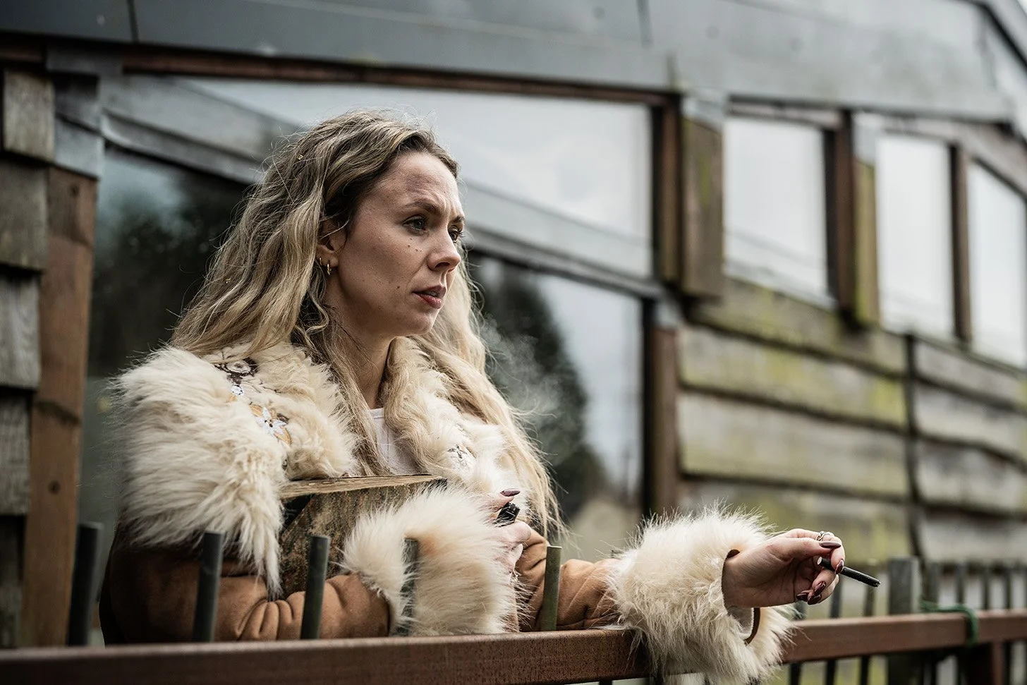 A woman with long blonde hair wearing a fur-trimmed jacket leaning on a balcony railing, holding a cigarette in her right hand, outdoors near a wooden building with large windows.