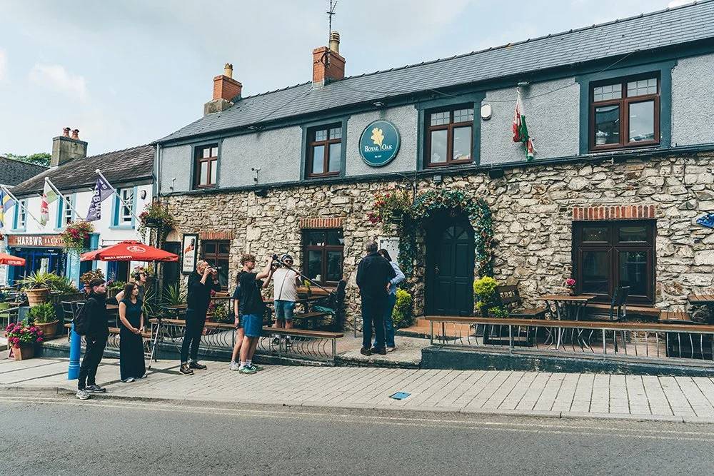 A film crew on the sidewalk filming outside a pub called Royal Oak, with people standing and talking in front of the pub.