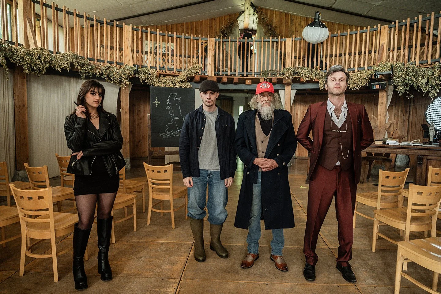 Four people standing in a rustic wooden room with chairs, a chalkboard with a map, and a balcony. The woman on the left is in a black leather jacket, black dress, and knee-high boots. The second person is in a cap, jeans, and boots. The third is an older man with a long beard, red cap, and a long coat. The fourth man is in a maroon suit with a white shirt and glasses hanging from his collar.