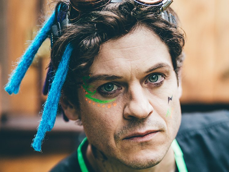 Close-up of a man with blue eyes and dark hair, wearing colorful face paint and a playful hairstyle with blue yarn in his hair. The background is blurred.