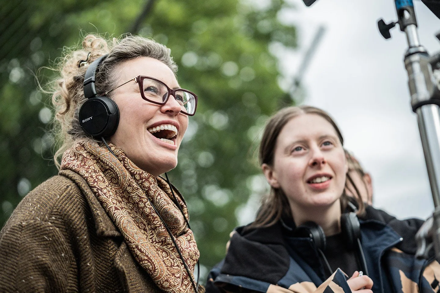 Two women outdoors, one smiling and wearing glasses and headphones, the other woman with headphones around her neck, looking at something off-camera, with trees in the background.