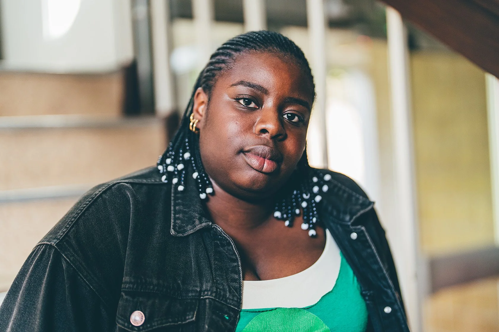 A young Black woman with braided hair decorated with white beads, wearing gold hoop earrings, a green and white top, and a black denim jacket, sitting on stairs indoors, looking at the camera.