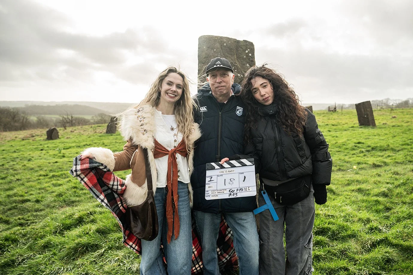 Three people standing outdoors on a cloudy day, with old stone structures in the background, possibly filming a scene or taking part in a historical re-enactment.