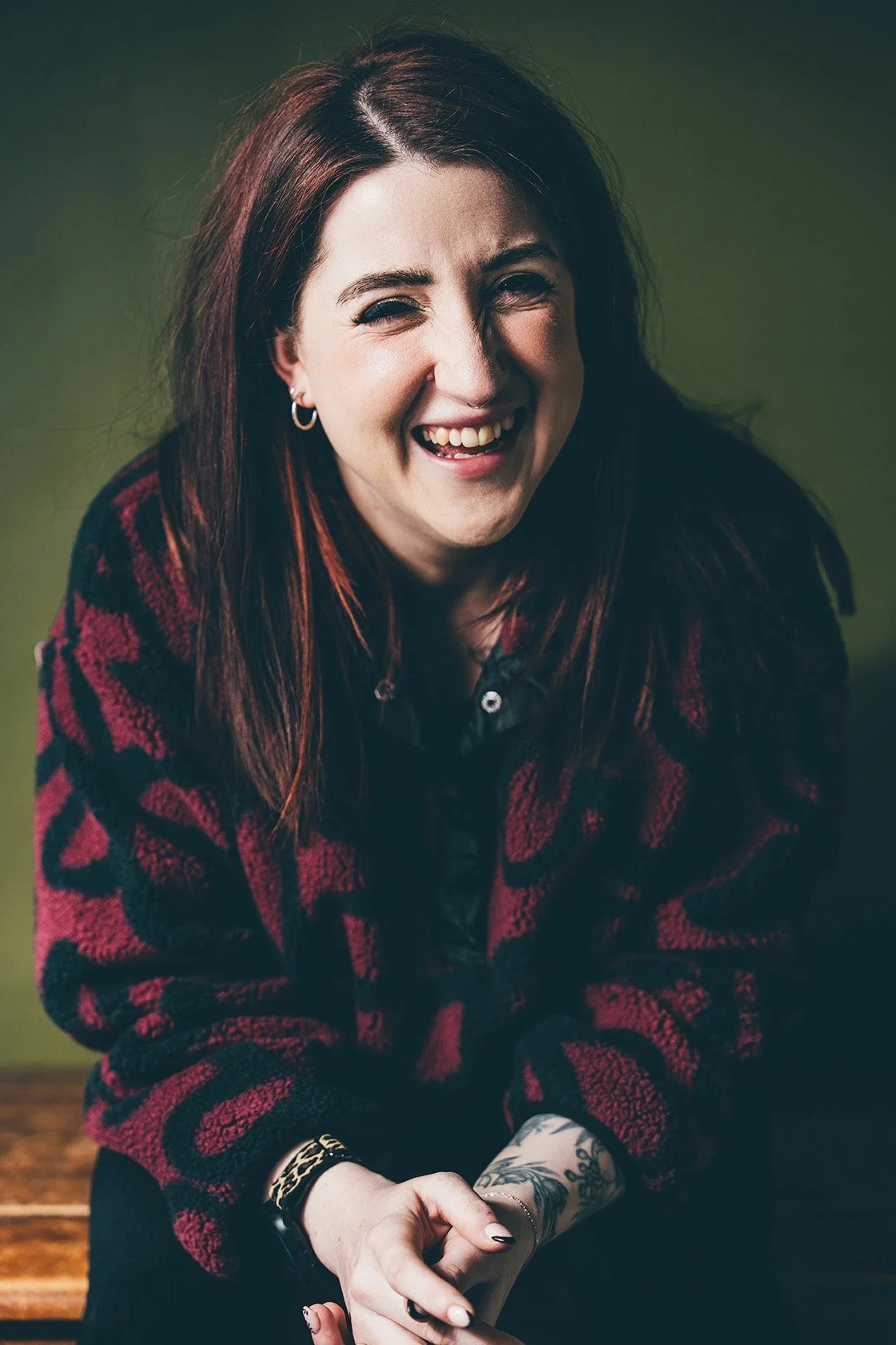 A woman with red hair laughing, wearing a red and black patterned sweater, with tattoos on her wrist and hand, sitting against a green background.