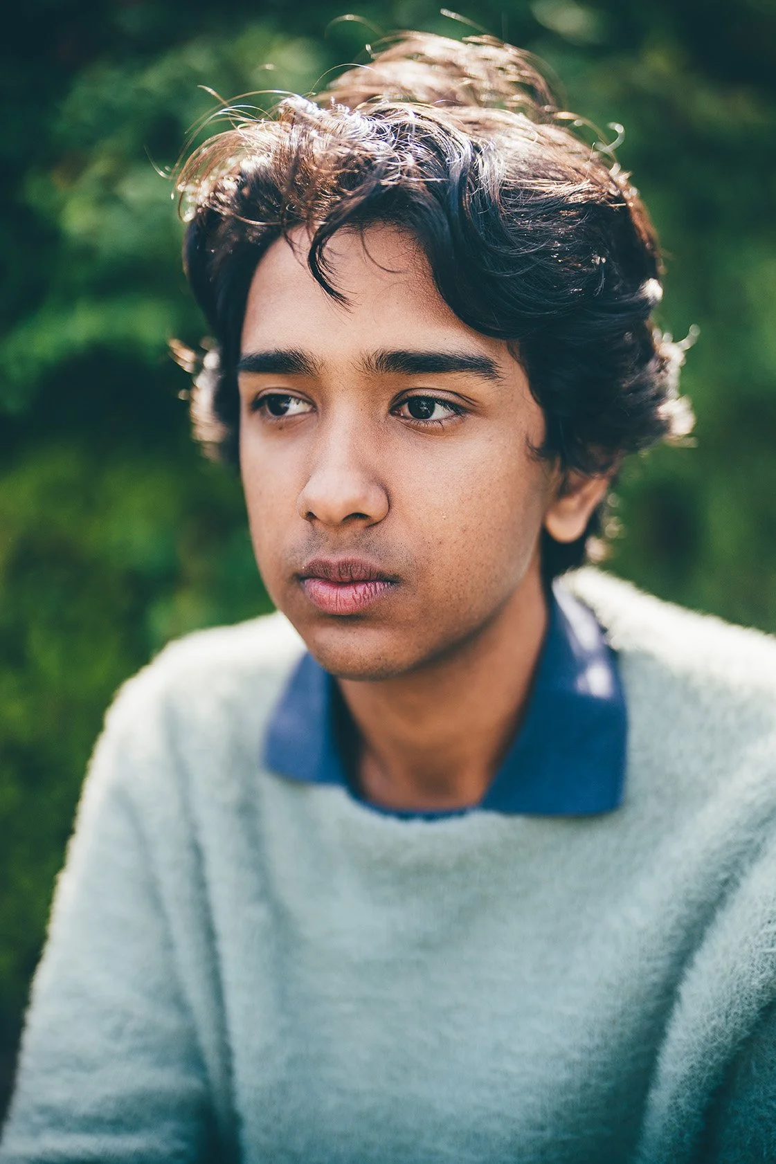 A young man with dark, wavy hair and medium skin tone is outdoors with a blurred green background, wearing a light-colored sweater over a blue collared shirt, looking off to the side with a neutral expression.