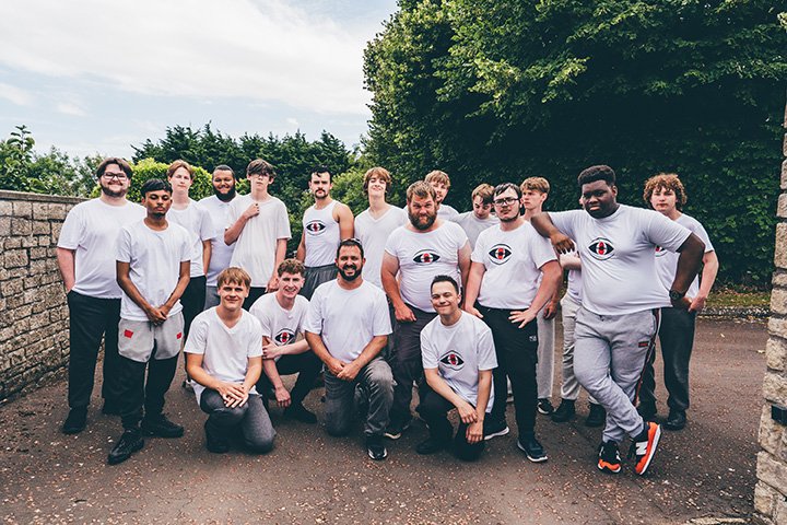 Group of young men and boys outdoors, some wearing matching white T-shirts with a logo, standing and sitting on a paved area with trees and a stone wall in the background.